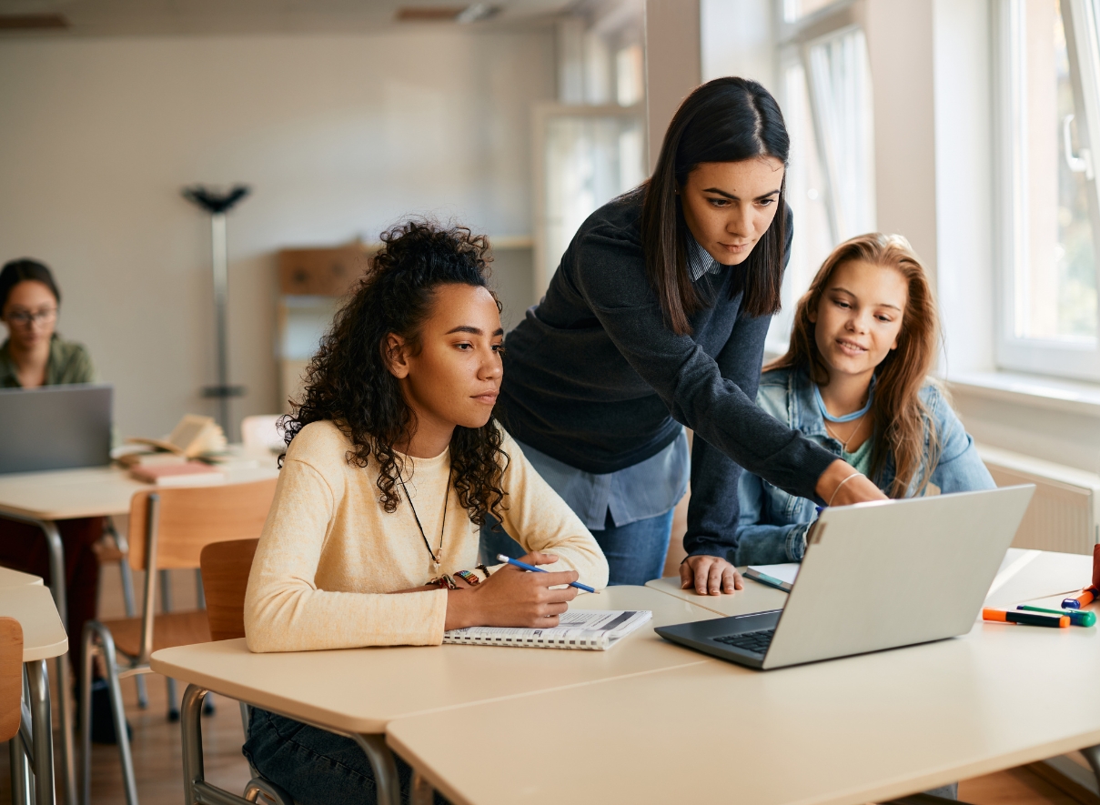 Male teacher pointing at a laptop. On his left side, a blonde female teenage student. On his right side, a black teenage student wearing glasses is typing in the laptop. To her right, a male, white, teenage student with a buzzcut. 